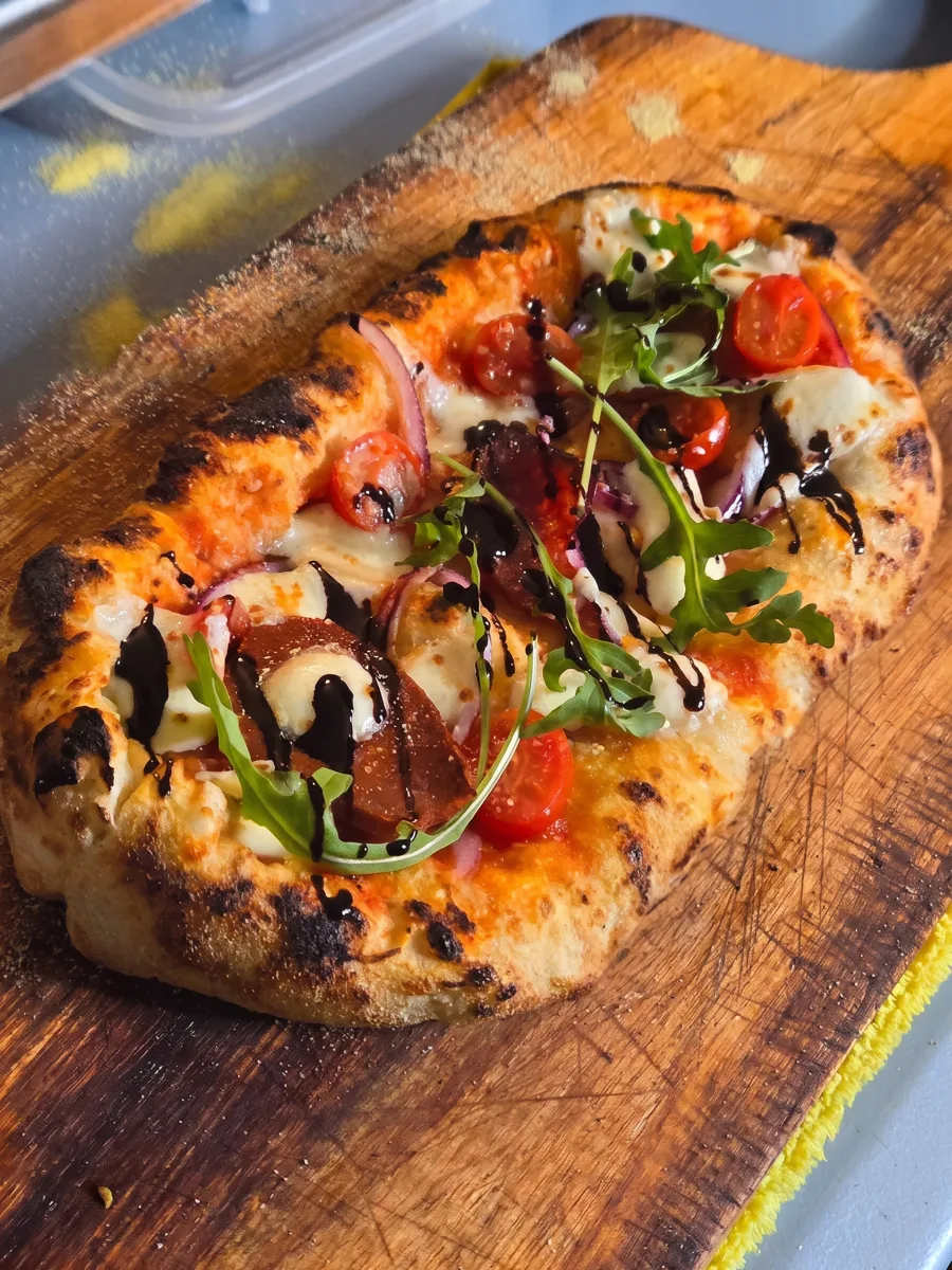 Pizza de La Ricetta recién salida, con tomates cherry, arúgula y glaseado balsámico, sobre su tabla